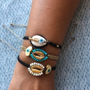 Puede incluir: Tres pulseras con cordón negro y detalles dorados. Cada pulsera presenta una concha blanca con un amuleto de ojo azul. Las pulseras están superpuestas en la muñeca de una persona.