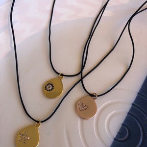 May include: Three necklaces on black cords featuring gold-toned teardrop-shaped pendants. One pendant displays a blue and white circular stone inlay, another features a small crystal-encrusted heart, and the third has a crystal-encrusted star.