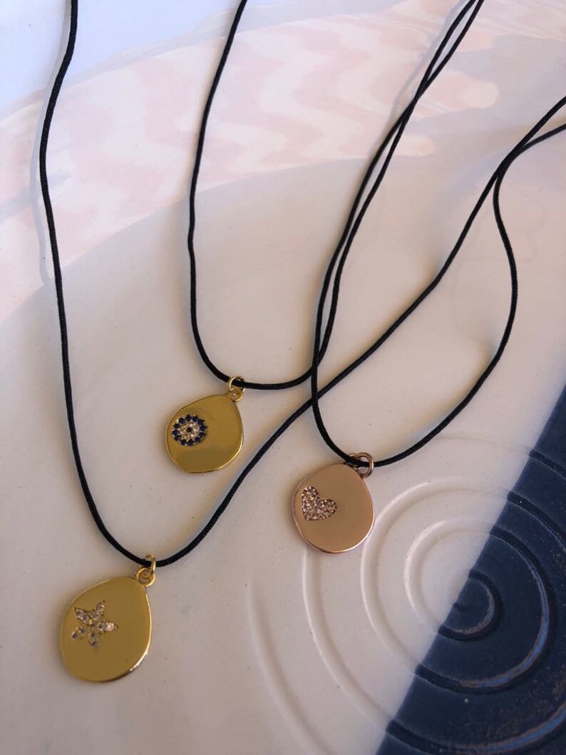 May include: Three necklaces on black cords featuring gold-toned teardrop-shaped pendants. One pendant displays a blue and white circular stone inlay, another features a small crystal-encrusted heart, and the third has a crystal-encrusted star.