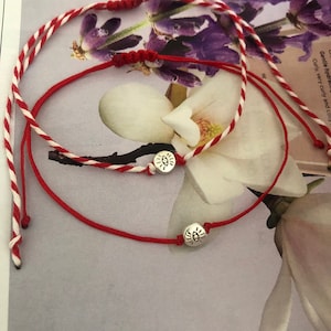 May include: Two red and white string bracelets with silver charms. The charms are round and have a simple eye design.