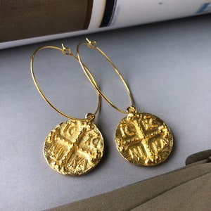 May include: Gold hoop earrings with a round coin charm featuring a cross and the text "IC XC NIKA".