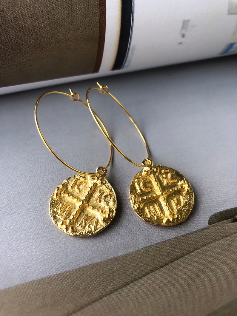May include: Gold hoop earrings with a round coin charm featuring a cross and the text "IC XC NIKA".