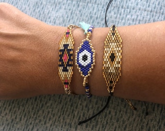 May include: Three beaded bracelets in gold, black, blue, and pink. The bracelets feature geometric and eye-shaped designs. One bracelet has a blue tassel. The bracelets are on a wrist.
