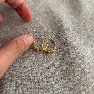 K&ouml;nnte beinhalten: Zwei kleine, goldfarbene Creolen mit kleinen, klaren Verzierungen. Die Ohrringe sind rund und scheinen aus Metall zu sein. Sie werden auf einer strukturierten, grauen Oberfl&auml;che gezeigt.