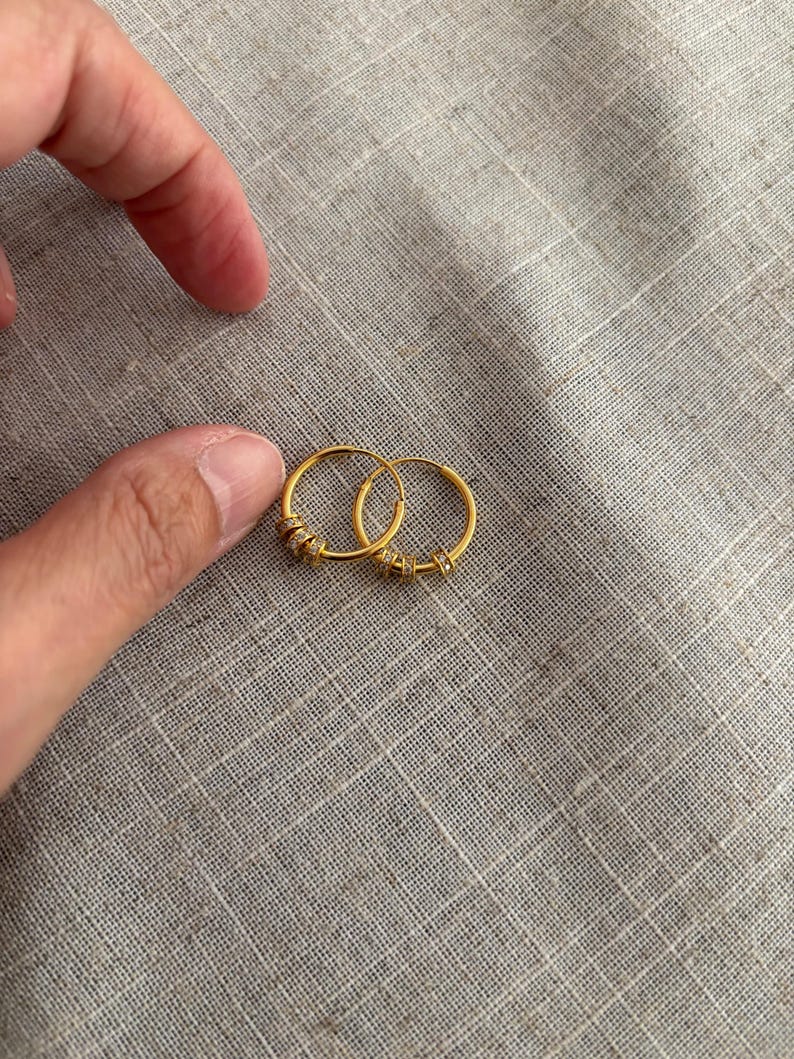 K&ouml;nnte beinhalten: Zwei kleine, goldfarbene Creolen mit kleinen, klaren Verzierungen. Die Ohrringe sind rund und scheinen aus Metall zu sein. Sie werden auf einer strukturierten, grauen Oberfl&auml;che gezeigt.