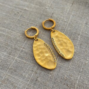 May include: A pair of gold-colored earrings. Each earring features a hammered, oval-shaped pendant with a row of small, clear stones along one edge. The earrings have a simple hoop closure. The background is a neutral, textured fabric.