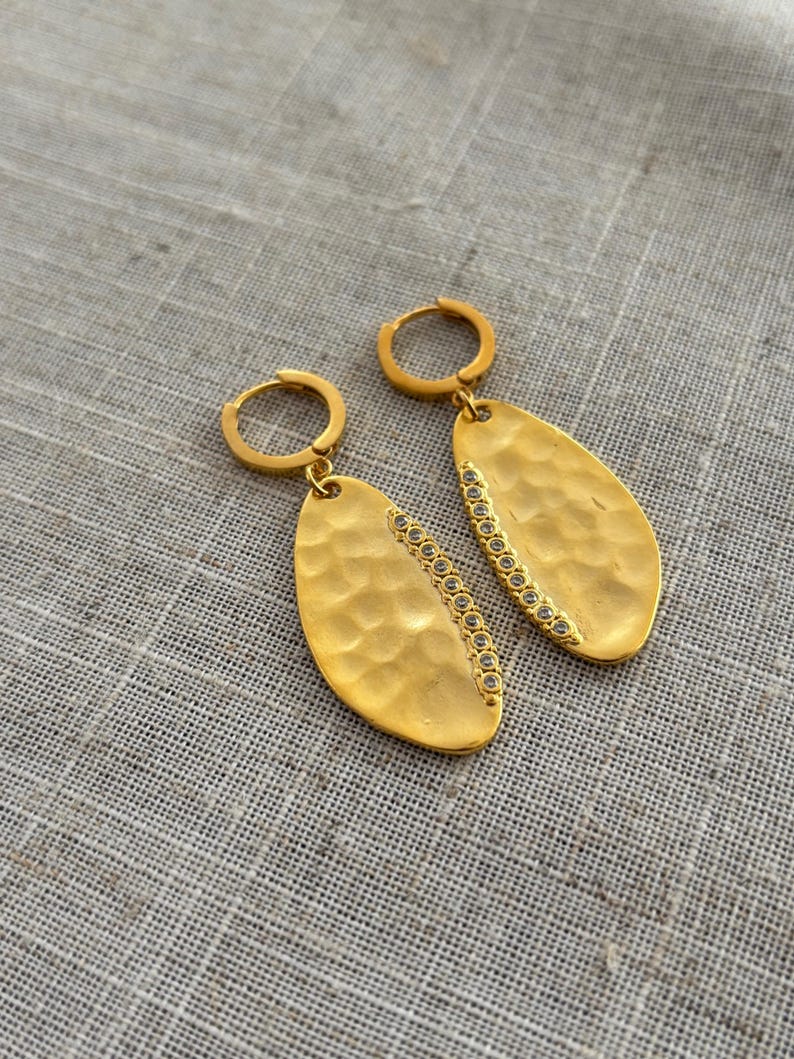 May include: A pair of gold-colored earrings. Each earring features a hammered, oval-shaped pendant with a row of small, clear stones along one edge. The earrings have a simple hoop closure. The background is a neutral, textured fabric.