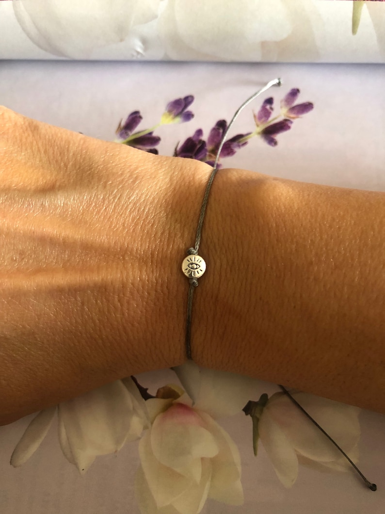 May include: A gray string bracelet with a silver charm featuring an eye symbol.