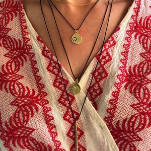 May include: A layered necklace set featuring a thin gold chain and three black cords. The cords hold gold-toned pendants: a circular charm with a small dark eye motif, a circular charm with a star engraving, and a long, slender gold-toned bar pendant.