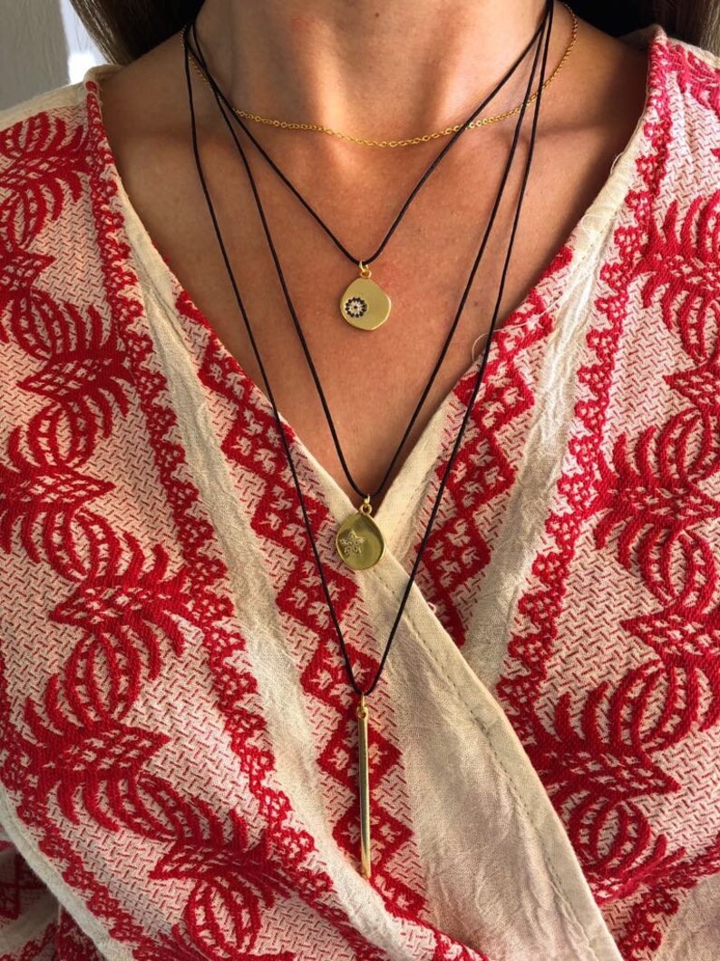 May include: A layered necklace set featuring a thin gold chain and three black cords. The cords hold gold-toned pendants: a circular charm with a small dark eye motif, a circular charm with a star engraving, and a long, slender gold-toned bar pendant.