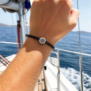 May include: A black nautical bracelet with a silver-colored ship's wheel charm. The bracelet is worn on a wrist, with a sailboat and ocean in the background. The bracelet is a simple, elegant accessory.