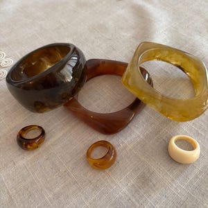 Può includere: Una collezione di gioielli in resina, tra cui tre braccialetti e tre anelli. I braccialetti sono nelle tonalità del marrone e dell'ambra, con un anello color crema. I gioielli sono esposti su un tessuto di colore neutro.