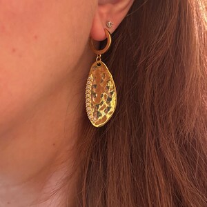 May include: Gold-toned hoop earring with a textured, teardrop-shaped pendant. The pendant is embellished with a row of small, sparkling stones along one edge. A small stud earring is also visible.