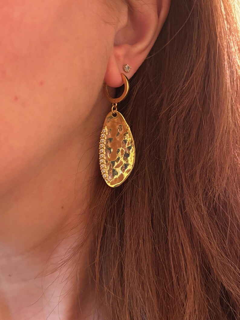 May include: Gold-toned hoop earring with a textured, teardrop-shaped pendant. The pendant is embellished with a row of small, sparkling stones along one edge. A small stud earring is also visible.