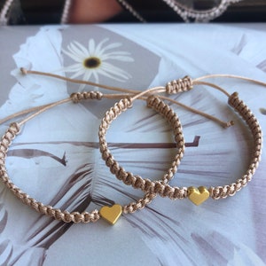 May include: Two beige friendship bracelets with gold heart charms. The bracelets are made of braided cord and have adjustable closures.