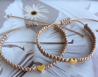 May include: Two beige friendship bracelets with gold heart charms. The bracelets are made of braided cord and have adjustable closures.