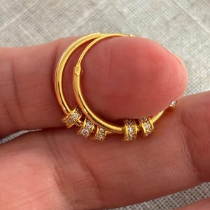 K&ouml;nnte beinhalten: Ein Paar goldfarbene Creolen. Jeder Ohrring hat mehrere kleine, klare Edelsteine, die in einer Reihe entlang der unteren H&auml;lfte angeordnet sind. Die Ohrringe werden in einer Hand gehalten.