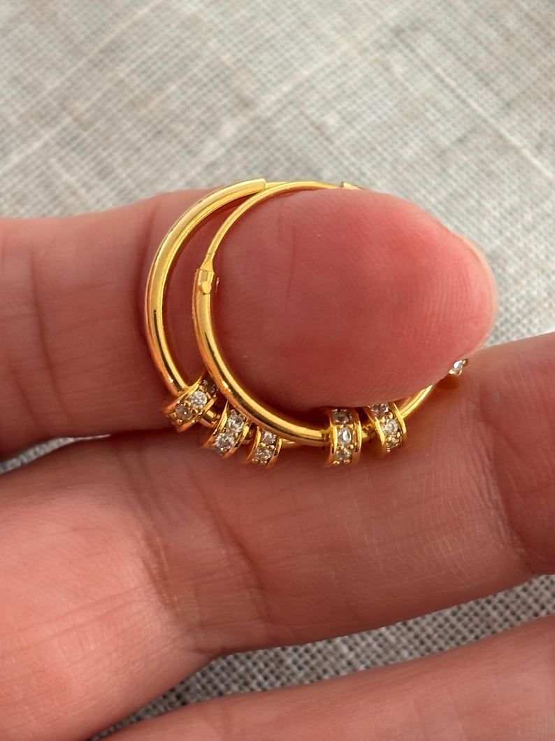 K&ouml;nnte beinhalten: Ein Paar goldfarbene Creolen. Jeder Ohrring hat mehrere kleine, klare Edelsteine, die in einer Reihe entlang der unteren H&auml;lfte angeordnet sind. Die Ohrringe werden in einer Hand gehalten.