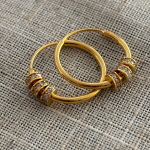 K&ouml;nnte beinhalten: Ein Paar goldener Creolen. Jeder Ohrring hat einen Abschnitt, der mit kleinen, klaren Edelsteinen verziert ist. Die Ohrringe werden auf einer strukturierten, neutralfarbenen Oberfl&auml;che gezeigt, die ihre polierte, reflektierende Oberfl&auml;che hervorhebt.