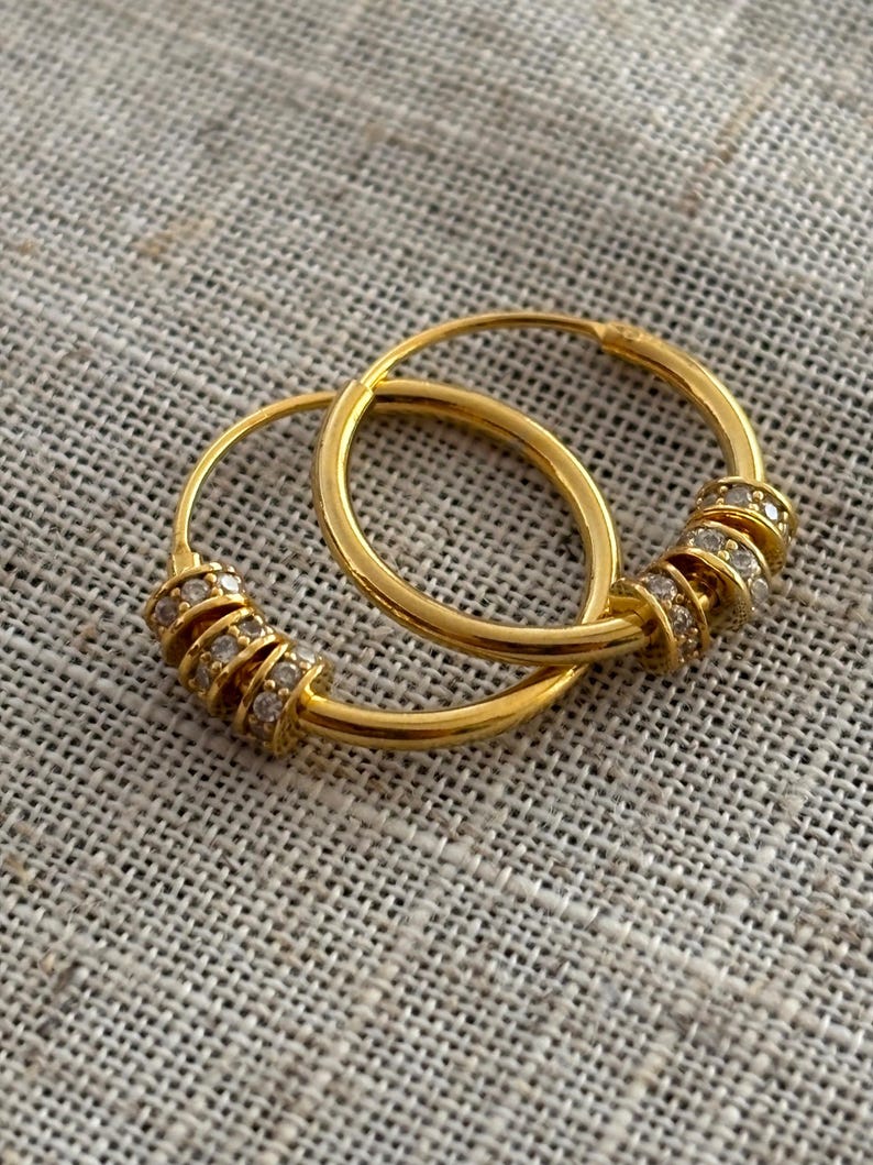 K&ouml;nnte beinhalten: Ein Paar goldener Creolen. Jeder Ohrring hat einen Abschnitt, der mit kleinen, klaren Edelsteinen verziert ist. Die Ohrringe werden auf einer strukturierten, neutralfarbenen Oberfl&auml;che gezeigt, die ihre polierte, reflektierende Oberfl&auml;che hervorhebt.