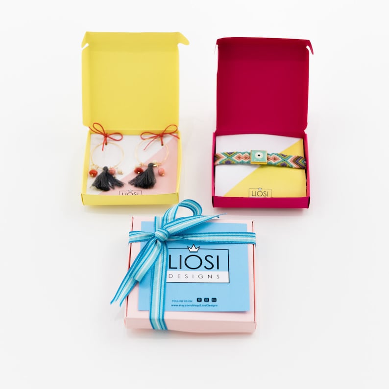 May include: Two open gift boxes containing jewelry: one yellow box with hoop earrings featuring dark gray tassels and red bows, and one magenta box with a woven bracelet featuring a square evil eye charm. A third closed pink box with a blue striped ribbon.