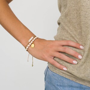 May include: A woman's wrist wearing two bracelets. One bracelet is a delicate gold chain with a small gold charm. The other bracelet is a string of white pearls.