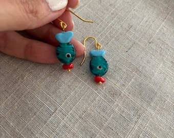 Puede incluir: Un par de pendientes colgantes con forma de pez, con cuerpos de cristal verde azulado con rayas oscuras, aletas de cristal azul claro y pequeñas cuentas de cristal rojo en la base. Cuentan con ganchos de metal dorado para orejas.