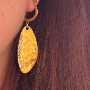 May include: A gold-colored earring with a hammered texture and a teardrop shape. The earring is adorned with small, clear gemstones along one edge. The earring is hanging from a gold hoop. The image shows the earring worn on an ear.