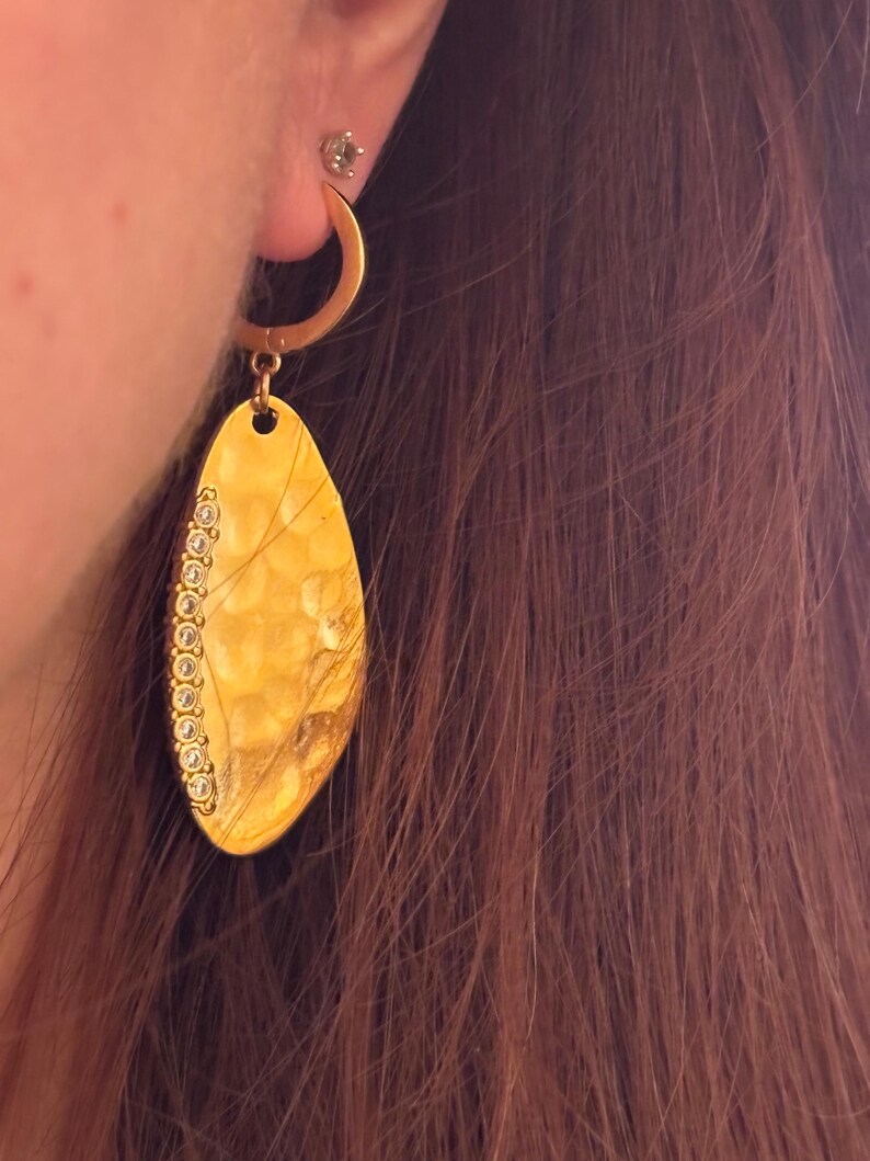 May include: A gold-colored earring with a hammered texture and a teardrop shape. The earring is adorned with small, clear gemstones along one edge. The earring is hanging from a gold hoop. The image shows the earring worn on an ear.