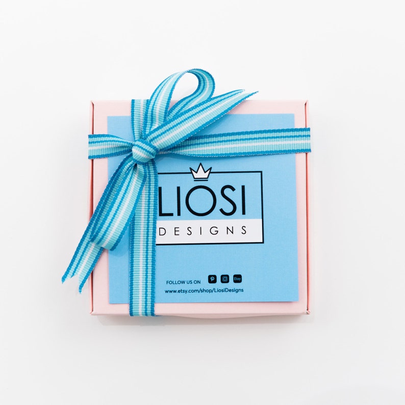May include: A pink gift box with a blue and white striped ribbon tied in a bow. The box has a blue label with the text "Liosi Designs" and the text "Follow us on" with social media icons and a website address.