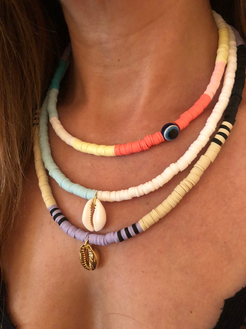 May include: A layered necklace with colorful beads and shell charms. The top layer features a blue evil eye charm. The necklace is made of white, light blue, yellow, pink, and black beads.