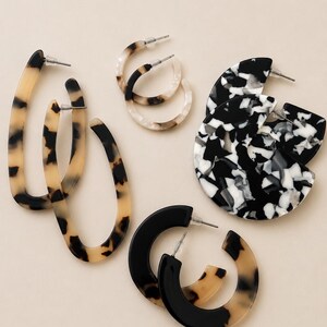 Op de afbeelding: Een collectie van vier paar oorbellen met diverse vormen en patronen. Inbegrepen zijn langwerpige ovale ringen in beige en zwart schildpadpatroon, kleine ronde ringen in crème en bruin, grote zwart-wit gemarmerde schijven en dikke zwarte en beige ringen.