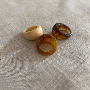 Op de afbeelding: Drie dikke harsringen in ivoor, amber en schildpadpatronen. De ringen zijn geplaatst tegen een neutrale linnen achtergrond. De ringen hebben een gladde, glanzende afwerking en een gedurfd, modern design, geschikt voor dagelijks gebruik.