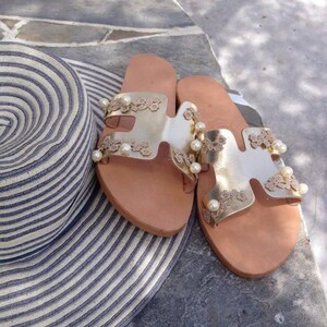 May include: A pair of gold leather sandals with delicate lace detailing and pearl accents. The sandals have a simple, strappy design and are perfect for summer.