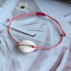Puede incluir: Un brazalete de cordón trenzado rojo y blanco con un dije de concha blanca.