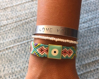 May include: A silver bracelet with the words "I LOVE YOU" engraved on it. The bracelet is stacked with a white pearl bracelet and a colorful woven bracelet with a turquoise evil eye charm.