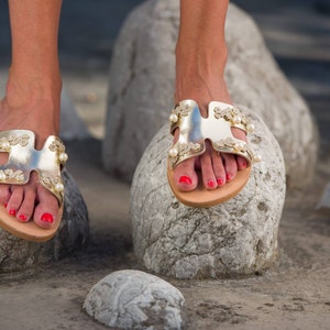 May include: Gold sandals with pearl embellishments and a strap design. The sandals are worn on a woman's feet and are resting on a rock.