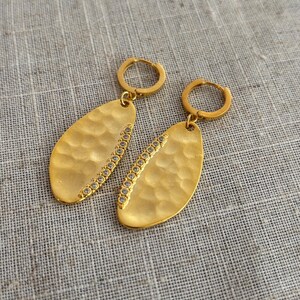 May include: A pair of gold-colored earrings with a hammered texture and a row of small, clear stones along one side. The earrings have a teardrop shape and are suspended from small hoop closures.