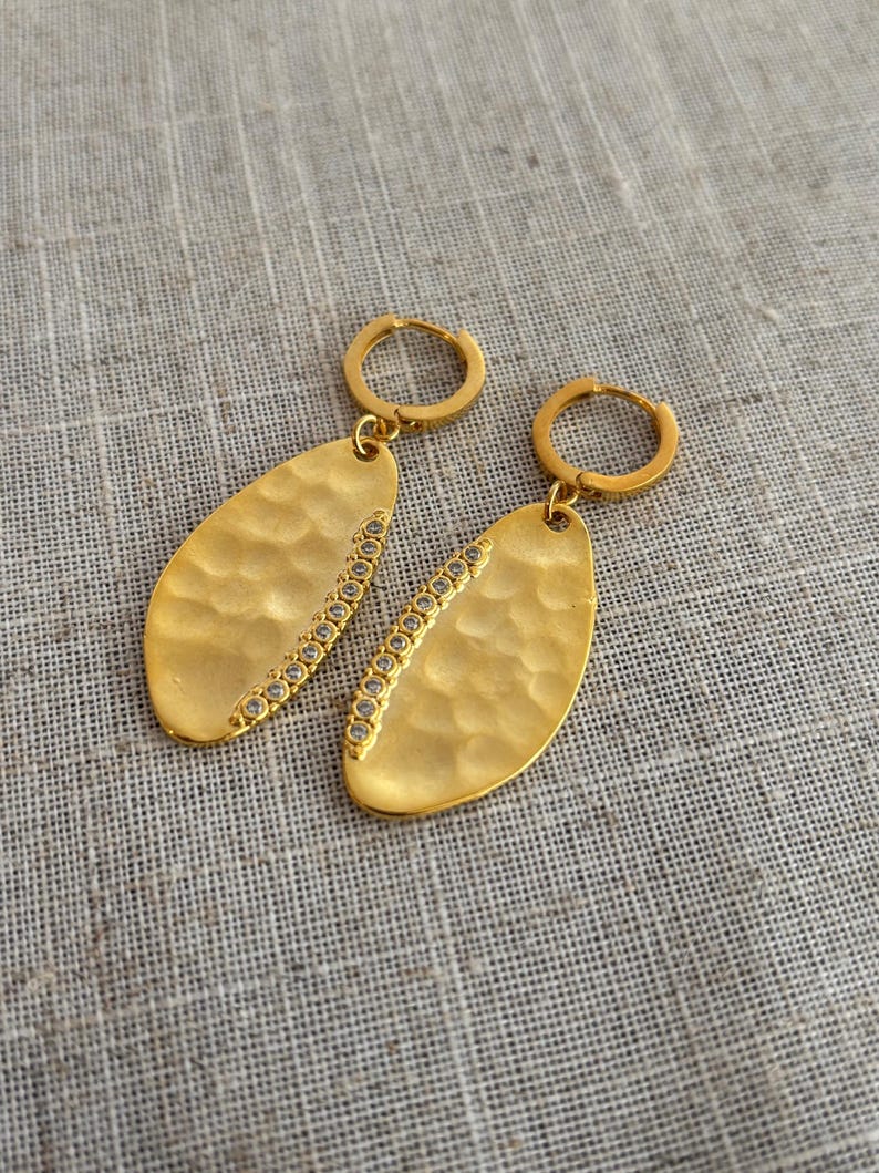 May include: A pair of gold-colored earrings with a hammered texture and a row of small, clear stones along one side. The earrings have a teardrop shape and are suspended from small hoop closures.