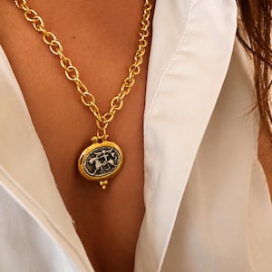 May include: A gold-toned chain necklace featuring a large oval pendant with a silver-coloured centre. The pendant displays an embossed figure riding an animal. The chain consists of thick, textured circular links, and the pendant has a small decorative bead at the bottom.