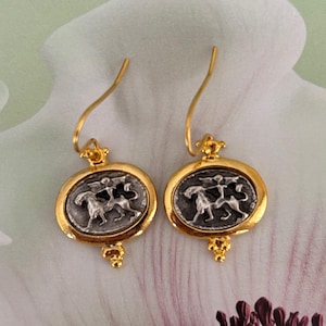 May include: A pair of gold earrings with a silver circular pendant featuring a detailed design of two horses and a rider. The earrings have a drop design with a small ball at the bottom.