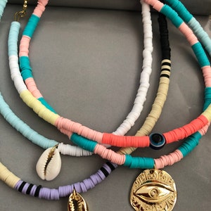 May include: Three layered necklaces with colorful beads and gold charms. The top necklace has pink, yellow, blue, and white beads. The middle necklace has black and white beads. The bottom necklace has purple beads. The charms are a gold seashell and a gold evil eye.