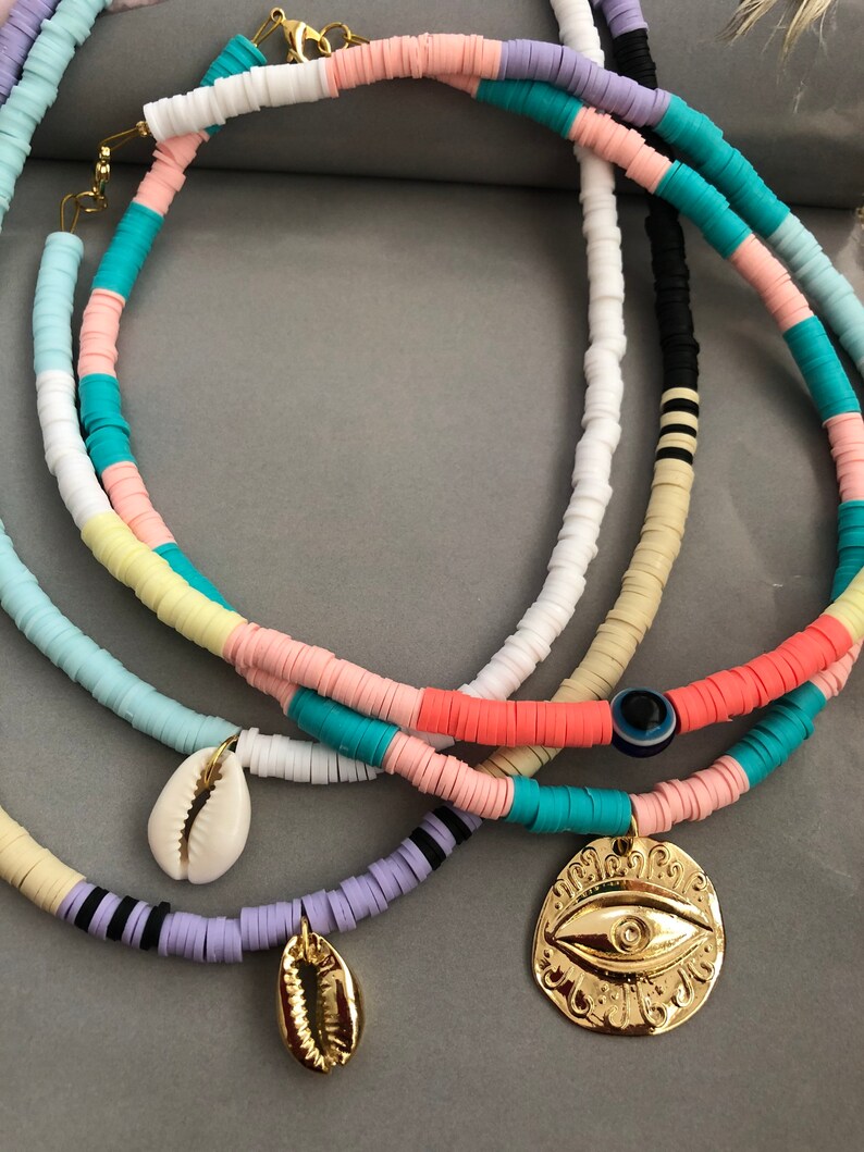 May include: Three layered necklaces with colorful beads and gold charms. The top necklace has pink, yellow, blue, and white beads. The middle necklace has black and white beads. The bottom necklace has purple beads. The charms are a gold seashell and a gold evil eye.