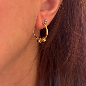 K&ouml;nnte beinhalten: Goldene Creolen mit drei kleinen, dekorativen Anh&auml;ngern. Die Anh&auml;nger scheinen mit kleinen, klaren Steinen besetzt zu sein. Ein kleiner, klarer Ohrstecker ist ebenfalls im Ohr zu sehen. Die Ohrringe sind goldfarben.