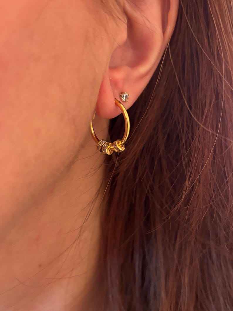 K&ouml;nnte beinhalten: Goldene Creolen mit drei kleinen, dekorativen Anh&auml;ngern. Die Anh&auml;nger scheinen mit kleinen, klaren Steinen besetzt zu sein. Ein kleiner, klarer Ohrstecker ist ebenfalls im Ohr zu sehen. Die Ohrringe sind goldfarben.