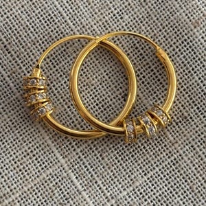 K&ouml;nnte beinhalten: Ein Paar goldene Creolen. Jeder Ohrring hat eine Reihe kleiner, klarer Edelsteine, die in einem Band um den Reifen angeordnet sind. Die Ohrringe werden auf einer strukturierten, neutralfarbenen Oberfl&auml;che pr&auml;sentiert.