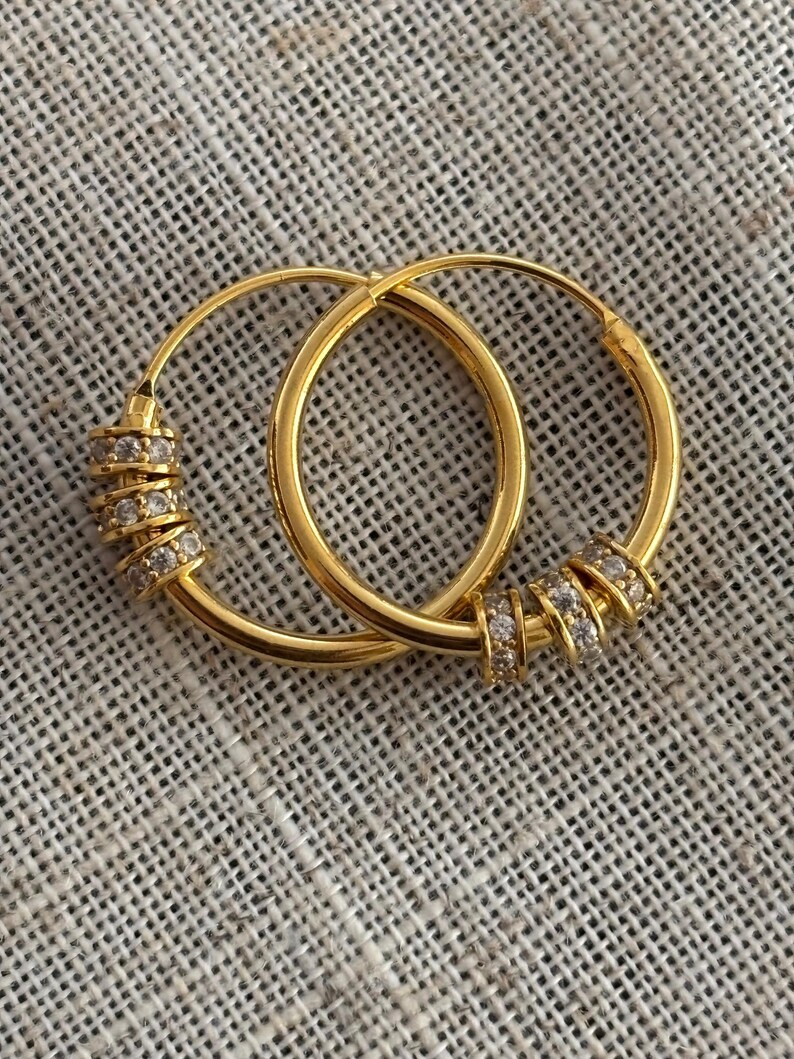K&ouml;nnte beinhalten: Ein Paar goldene Creolen. Jeder Ohrring hat eine Reihe kleiner, klarer Edelsteine, die in einem Band um den Reifen angeordnet sind. Die Ohrringe werden auf einer strukturierten, neutralfarbenen Oberfl&auml;che pr&auml;sentiert.