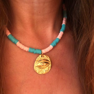 May include: A gold necklace with a large, ornate eye charm. The necklace is made of pink and turquoise beads.