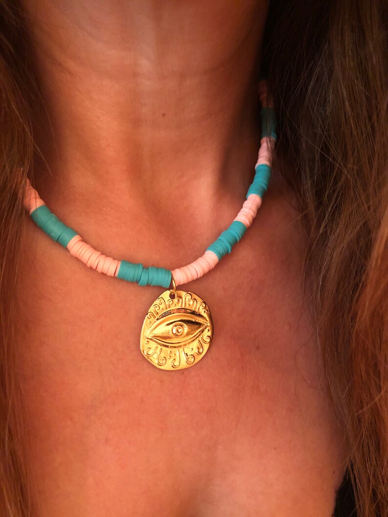May include: A gold necklace with a large, ornate eye charm. The necklace is made of pink and turquoise beads.