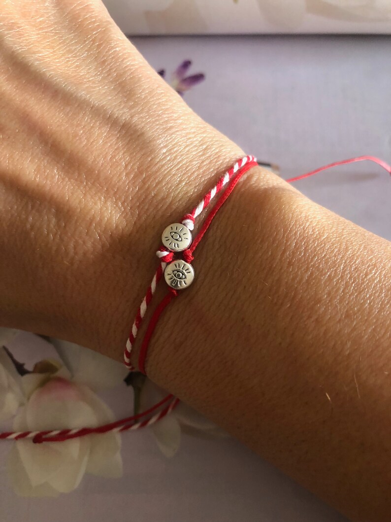May include: Red and white string bracelet with two silver charms featuring an eye symbol.